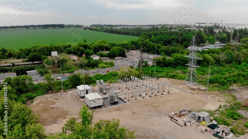 Aerial drone view of an electrical substation under construction in Sumy, Ukraine. High-voltage transformers, switchgear, transmission equipment, utility buildings, workers, and a yellow excavator