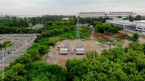 Aerial drone view of an electrical substation under construction in Sumy, Ukraine. High-voltage transformers, switchgear, transmission equipment, utility buildings, workers, and a yellow excavator
