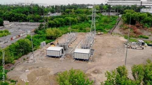Aerial drone view of an electrical substation under construction in Sumy, Ukraine. High-voltage transformers, switchgear, transmission equipment, utility buildings, workers, and a yellow excavator