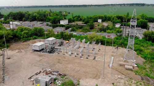 Aerial drone view of an electrical substation under construction in Sumy, Ukraine. High-voltage transformers, switchgear, transmission equipment, utility buildings, workers, and a yellow excavator