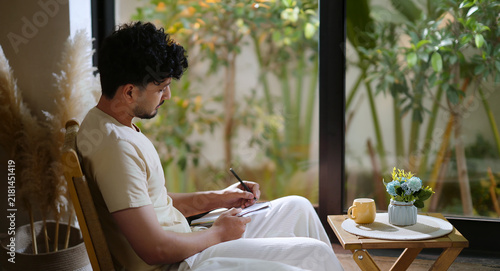 Indian calm young adult gen z man sitting on chair enjoy day resting indoor room. Male boy hold pen writing book new work notes plan ideas spend time take casual job break home house place