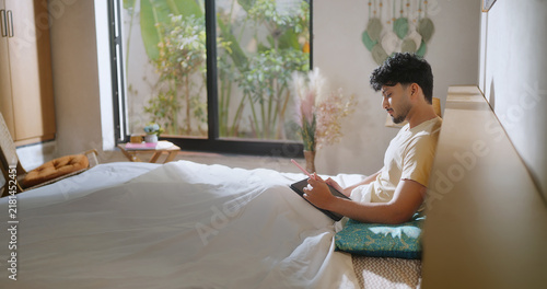 Indian young adult gen z male man sitting on bed use tab look screen do study work check email boy hold tablet stylus pen writing plan new notes ideas spend time take job break room home place