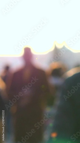 Backlit crowd gathers outdoors in warm golden light. Soft blurred figures stand close together while sun glare silhouettes heads and shoulders against bright sky and distant tents.
