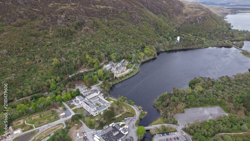 Aerial view of Kylemore Abbey nestled beside the dark waters of Pollacappul Lough, surrounded by lush green forests and rugged mountains in Connemara, County Galway, Ireland.