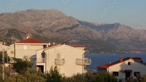 Sunlit coastal houses beneath dramatic mountains beside calm blue sea