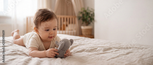 A happy baby lies on a comfortable bed holding a plush animal toy in a bright sunny bedroom. cheerful morning relaxation, innocent child moment, cozy home, header banner topbar.