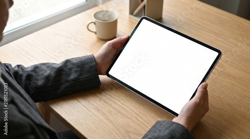 Person holding modern digital tablet with empty blank white screen while working at wooden desk