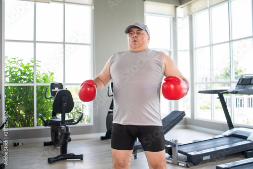 Fotografie An overweight mature man wearing red boxing gloves stands inside a bright modern gym with large windows, training for fitness, health, and weight loss with determination and focus