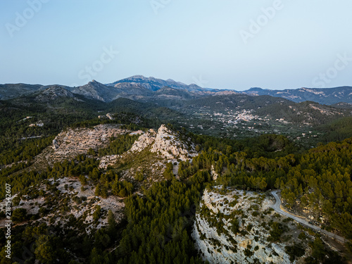 Mallorca, Spain - 15 April 2024: Aerial view of the rugged landscape of the Serra de Tramuntana mountains with dense pine forests, rocky cliffs, and a winding road under a clear sky.