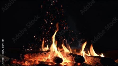 A low intense fire glowing in complete darkness with hundreds of tiny orange embers slowly floating upward. The main flames remain concentrated near the bottom center while sparks travel through the s
