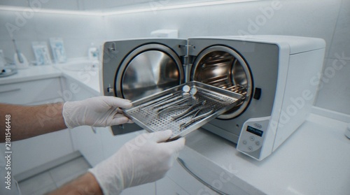 Bild auf Leinwand A dentist wearing white gloves is placing a tray of sterilized metal dental instruments into a steam autoclave machine in a modern dental clinic