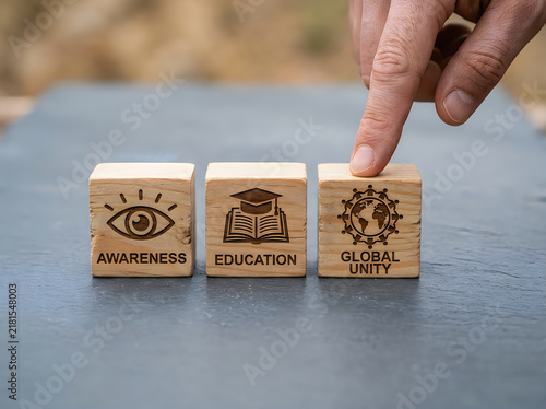 Wooden blocks featuring engraved icons of awareness education and global unity placed together by a person on gray stone surface