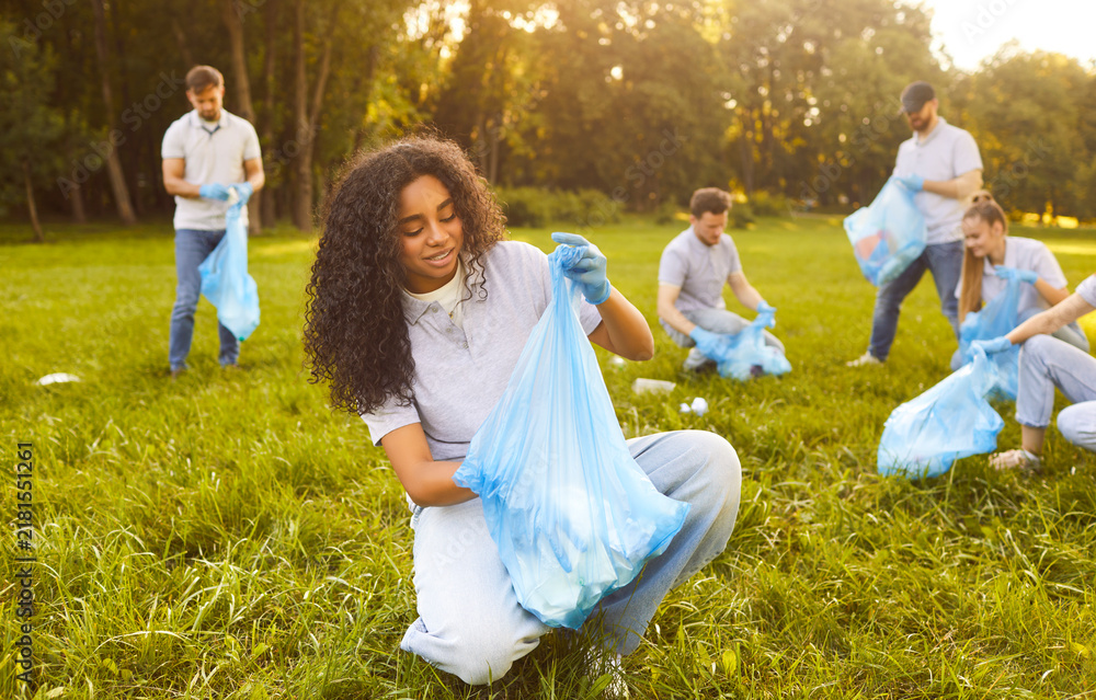 Obraz premium Happy young people volunteers with trash bags collecting garbage from green grass in summer park. Men and women cleaning trash in nature. Environmental pollution, volunteering and ecology concept