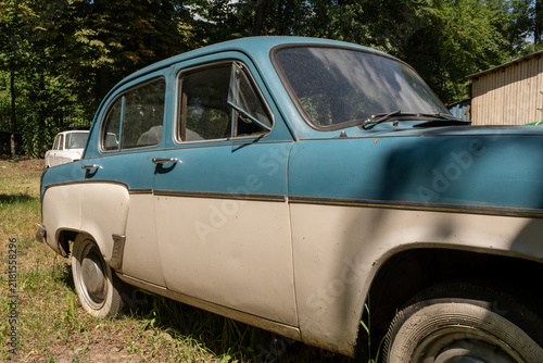 Картината върху платно Classic blue and white retro sedan with weathered body and whitewall tires stands on dry grass near trees