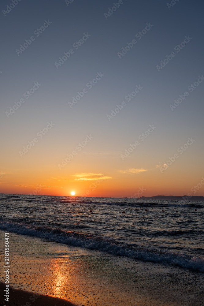 Obraz premium Sun touches sea horizon casting orange light on rippling waves. Coastal view with wet beach shore, sunset reflection on sand, gradient sky, and distant island hills.