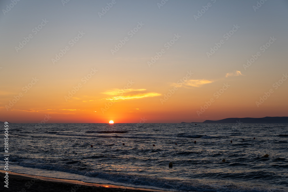 Obraz premium Sun touches sea horizon casting orange light on dark wavy water. Coastline view with sandy beach shore, small clouds in twilight sky, and distant island silhouette.