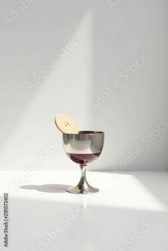 A solitary silver goblet, partially filled with a dark liquid, rests on a pristine white surface, a single round cracker delicately perched on its rim, bathed in soft, natural light.