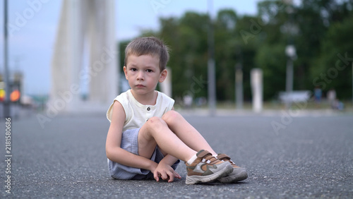 Foto Little sad boy sitting on asphalt pavement outdoors, legs tucked, serious face showing melancholy and isolation in quiet summer street scene