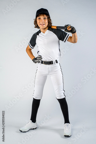 Male youth baseball player in full uniform, standing and smiling with bat over shoulder and hand on hip