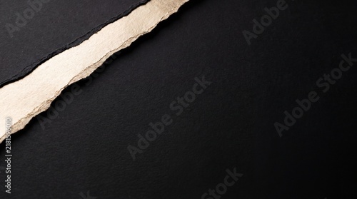 A torn edge of textured paper reveals a stark contrast against a deep black background, creating a minimalist and abstract composition with a strong sense of visual drama and tactile quality.