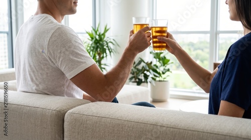 Happy couple toasting with beer glasses on sofa at home, casual lifestyle moment, man and woman relaxing in living room, celebration or date night, side view, bright natural