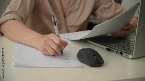 Close up footage of a woman writing notes on lined paper while consulting documents beside a laptop and wireless mouse at an office desk. Everyday planning, paperwork and administrative work.
