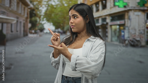 Принти на полотні Woman points index finger to the right with open palm held beneath, wearing a casual shirt and jeans on a pedestrian street by a pharmacy storefront; curiosity