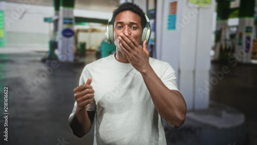 Fotografía Black man wearing headphones dancing and smiling with hands raised at a street gas station; joy music energy