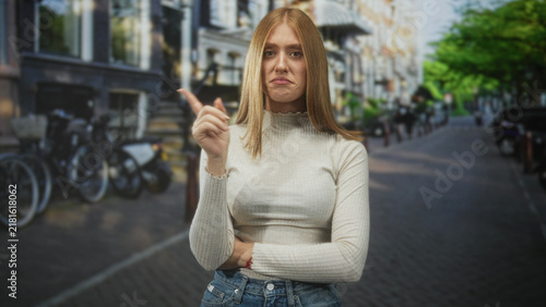 фотографія Woman points finger with crossed arm on cobblestone street beside parked bicycles and brick buildings; disapproval boundary assertion