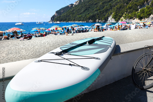 Inflatable stand up paddleboard with paddle resting on beach wall. Inflatable SUP board on sunny summer day at crowded sea beach resort. Active water sports equipment for outdoor recreation and summer