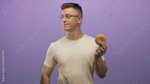 Bild auf Leinwand Man holds glazed donut up in studio while looking at it, wearing glasses and beige t shirt; craving contentment