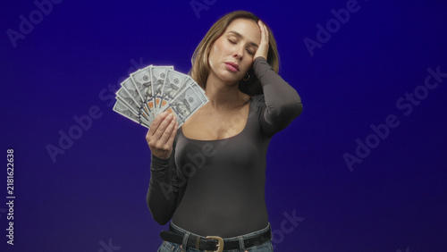 Cuadro en lienzo Woman holding a fan of hundred dollar bills with hand to temple, closed eyes and pained expression, wearing fitted top and jeans in studio; financial stress