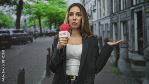Bild auf Leinwand Woman reporter in blazer holds red microphone labeled news, shrugs with one hand amid cobblestone city street and parked cars; uncertainty live update