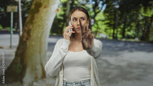 Fotografía Woman on a mobile phone points finger toward camera on a street beside a bus stop and park trees; assertive conversation