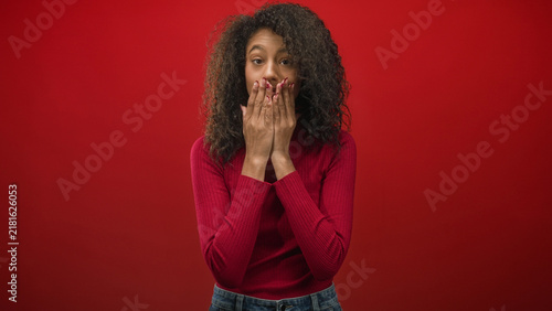 Fotografie Woman covers mouth with hands in studio with red backdrop and red sweater, curly hair and denim jeans; surprise delight