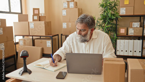 Obraz na plátně Man with gray beard writing notes at a laptop among parcel boxes in a packing area inside a building; diligence entrepreneurship