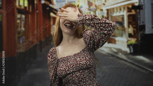 Quadro su tela Redhead woman laughing and waving hand on a narrow cobbled street with shopfronts and market pedestrians; joy carefree playfulness
