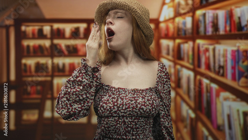 фотографія Redhead woman yawning with hand over mouth among bookstore shelves in a building, wearing floral dress and straw hat; sleepy boredom