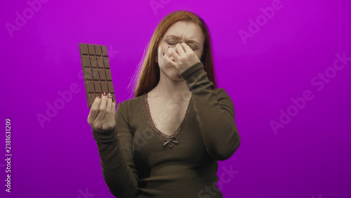 Obraz na płótnie Redhead woman holding chocolate bar, pinches nose and covers face against purple studio backdrop; disgust aversion