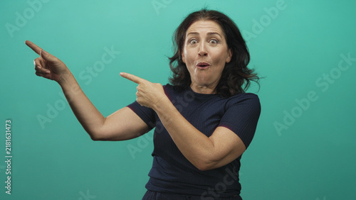 Fotografie Woman pointing index fingers with surprised open mouth and animated expression in studio teal backdrop; playful surprise