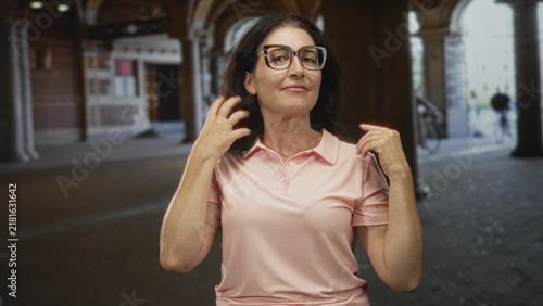 Konstfotografi Woman wearing pink polo adjusts hair and touches collar with hands under arched street arcade; confidence poise