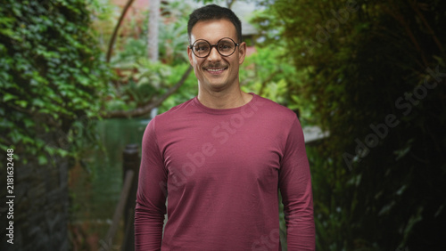 Hispanic man with glasses and moustache smiling and standing with arms relaxed in a lush garden courtyard wearing a maroon long sleeve shirt; serenity.