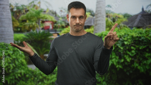 Foto Hispanic man displays index finger and open palm beside a tropical building, shrugging expression; dry humor doubt
