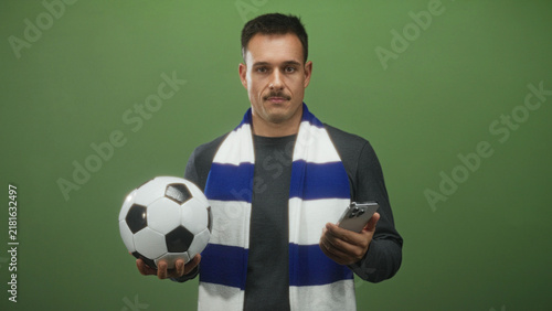 Fotografía Man holding soccerball in left hand and checking smartphone with right hand in studio, wearing blue and white fan scarf and moustache; team support anticipation