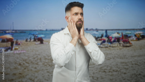 Obraz na płótnie Man covering ears with hands, pressing temples while wincing in a white shirt on a beach, suggesting a headache; anxiety overwhelm