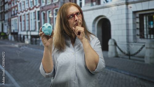 Fotografie Senior woman holds blue piggy bank and presses finger to lips on a cobblestone street by a stone building; secrecy savings caution
