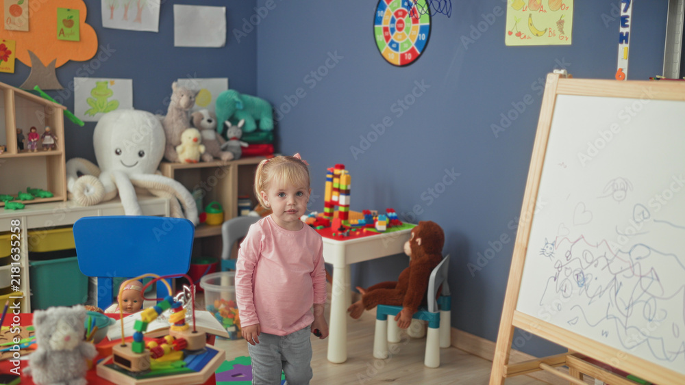 Fototapeta premium Little blonde girl stacks colorful blocks at a plastic play table amid plush toys and shelving in a preschool building; curiosity wonder imagination play.