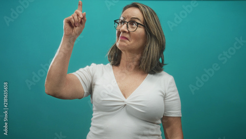 Obraz na płótnie Hispanic senior woman points finger up at an unseen spot, wearing glasses and white shirt against a teal studio backdrop; curiosity