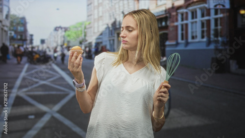 Foto Woman holding muffin in left hand and kitchen whisk in right, frowning and grimacing at the pastry while standing on a busy street lined with buildings; disgust doubt