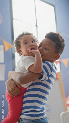 Bild auf Leinwand Older brother holds baby sister in his arms by a tall window and colorful bunting in a playroom building; affection